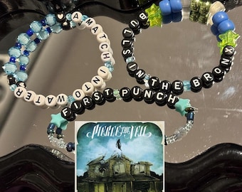 May include: Three beaded bracelets with blue and green beads and white letters spelling out "A Match Into First Punch In The Bronx". The bracelets are on a mirrored surface with a CD for the album "Collide With The Sky" by Pierce The Veil.