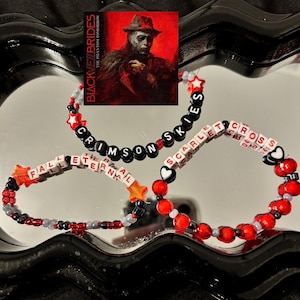 May include: Three beaded bracelets with the phrases "Crimson Skies", "Scarlet Cross", and "Fall Eternal" spelled out in letter beads. The bracelets are red, black, and white, with star and heart accents. A red album cover is in the background.