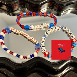 May include: Three beaded bracelets in red, white, and blue. The bracelets spell out "RITUAL", "BRAINWASHED", and "SUPER DARK" with white letter beads. A small red album cover with a blue frog graphic is also present.