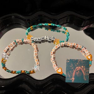May include: Three beaded bracelets with text and charms. One bracelet reads "Dizzy on the meadow", another reads "Humming", and the third reads "Cutting my fingers off". The bracelets are teal, orange, and white.
