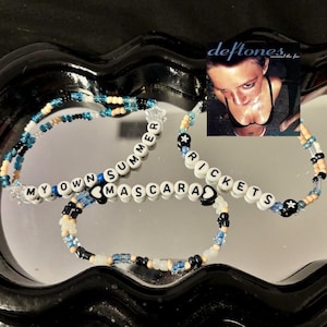 May include: Three beaded bracelets with white letter beads spelling "MY OWN SUMMER", "MASCARA", and "RICKETTS". The bracelets are accented with black, blue, and tan beads. A small image of a woman is in the background.