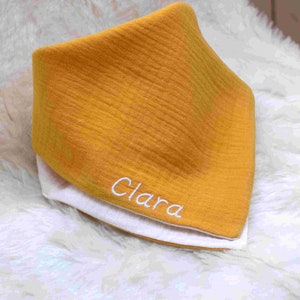 Pode incluir: Um babador de bebé em gaze de algodão amarelo mostarda com costuras brancas e o nome "Clara" bordado em fio branco.