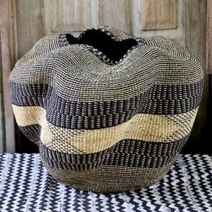 May include: A large, woven basket with a unique, wavy shape. The basket features alternating bands of black and natural beige fibers, creating a striking visual contrast. The open top reveals a dark interior, and the basket sits on a patterned surface.