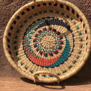 Pode incluir: Uma cesta redonda tecida com duas alças. A cesta é feita de materiais naturais e tem um padrão geométrico colorido em tons de azul, verde, laranja e preto.