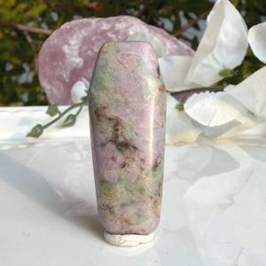 May include: A polished, coffin-shaped stone carving featuring a mottled surface with blended hues of soft pink, pale green, and dark brown. The object has a smooth, reflective texture and stands upright on a small, light-colored base.
