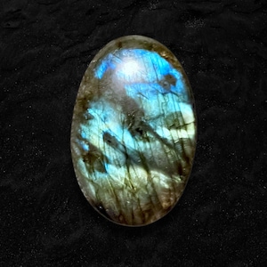 May include: Oval-shaped labradorite gemstone with a polished surface. The stone displays a mesmerizing play of color, with iridescent flashes of blue, green, and gold. The gemstone is set against a dark, textured background, highlighting its natural beauty.