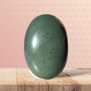 May include: A smooth, oval-shaped, teal-colored decorative egg with small dark spots. The egg is displayed on a wooden surface against a pink background with a dotted pattern. The egg has a glossy finish.
