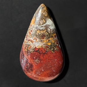 Könnte beinhalten: Ein tropfenförmiger, polierter Edelstein. Der Stein zeigt eine Mischung aus Farben, darunter tiefe Rottöne, Braun, Gelb und Weiß, die ein einzigartiges Muster bilden. Der Edelstein ist vor einem dunklen Hintergrund platziert.