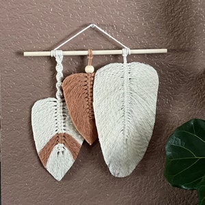 May include: A macrame wall hanging featuring three leaf-shaped designs in white and brown. The leaves are suspended from a wooden dowel with a white cord and a wooden bead. The background is a solid brown color.