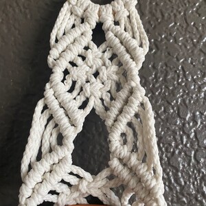 May include: A white macrame plant hanger with a wooden ring at the top. The hanger is designed to hold a small pot.