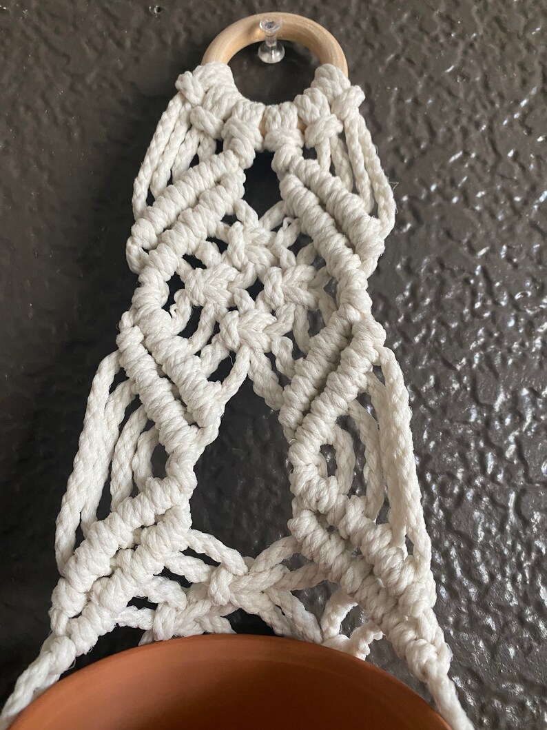 May include: A white macrame plant hanger with a wooden ring at the top. The hanger is designed to hold a small pot.