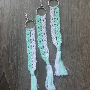 May include: Three macrame keychains with a white and light green color pattern. The keychains are made with a knotted cord and have a silver key ring.