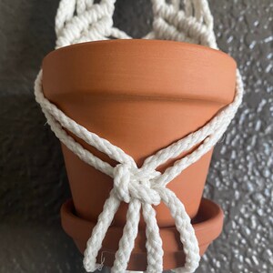 May include: A white macrame plant hanger with a terracotta pot hanging from it. The pot is empty and the hanger is made of thick rope.