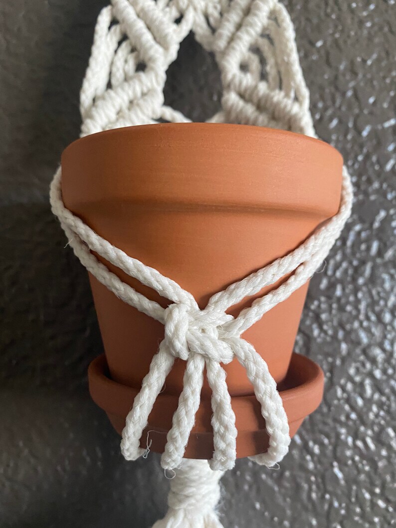 May include: A white macrame plant hanger with a terracotta pot hanging from it. The pot is empty and the hanger is made of thick rope.
