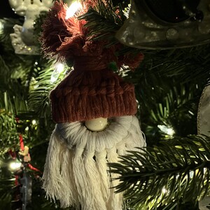 May include: A handmade gnome ornament with a red knitted hat, white beard, and a white bead for a nose. The ornament is hanging on a Christmas tree.