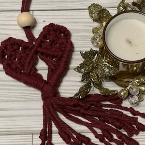May include: A burgundy heart-shaped macrame wall hanging with a tassel fringe. The hanging is made with thick cord and has a wooden bead at the top.
