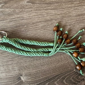 May include: Two keychains with silver rings and mint green macrame cords. Each cord is adorned with dark brown wooden beads. The keychains are displayed on a wooden surface.