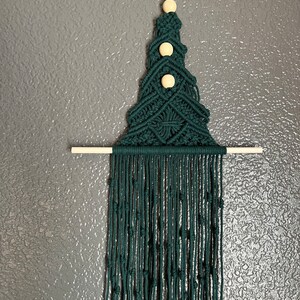 May include: A dark green macrame wall hanging in the shape of a Christmas tree with three wooden beads. The hanging has a fringe of tassels at the bottom.
