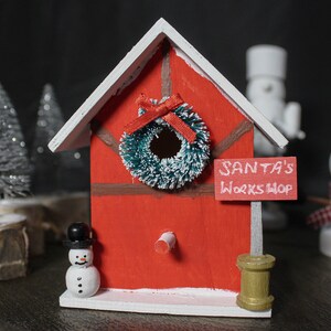 Può includere: Una casetta per uccelli rossa in legno del laboratorio di Babbo Natale con tetto bianco, ghirlanda verde e statuina di pupazzo di neve. Il cartello recita "SANTA'S WORKSHOP". Alberi di Natale argentati decorativi e un pupazzo di neve sullo sfondo.