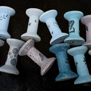 May include: A collection of eleven wooden spools painted in various shades of blue, gray, and lavender. Each spool features unique black stamped designs including botanical motifs, stars, buttons, letters, and an ampersand symbol on the surface.