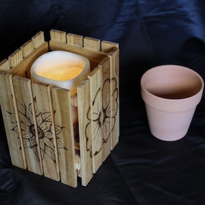 May include: A wooden candle holder with a sunflower design, and a terracotta flower pot. The candle holder is made of light-colored wood and has a square shape. The candle inside is lit, emitting a warm glow.