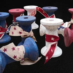 Op de afbeelding: Een verzameling houten klossen geschilderd in rood, wit en blauw, elk omwikkeld met stoffen lint met sterpatronen. De klossen zijn in een groep gerangschikt, waarbij sommige rechtop staan en andere op hun kant liggen, met een verweerde afwerking.