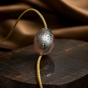 May include: A silver-colored, textured pendant with a gold-colored chain. The pendant is oval-shaped and hangs from a delicate, braided chain. The background is a blurred brown fabric.