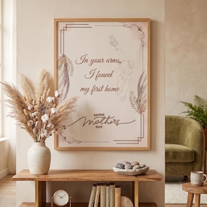 Peut inclure: Une affiche encadrée avec le texte "In your arms, I found my first home" et "Happy Mother's Day". L'affiche a un fond clair avec du texte marron et des éléments décoratifs. Un vase de fleurs séchées et une table en bois sont au premier plan.