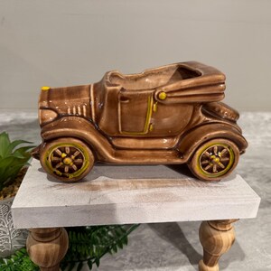 Puede incluir: Un macetero de cerámica marrón con forma de coche antiguo. El coche tiene detalles amarillos en las ruedas y los adornos. El macetero está sobre un soporte de madera blanco.