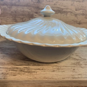Może przedstawiać: Biała ceramiczna miska do serwowania z pokrywką. Miska ma falisty brzeg, a pokrywka ma żebrowany wzór. Miska stoi na drewnianej powierzchni.