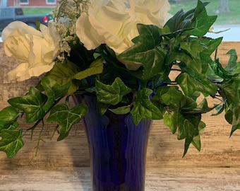 May include: A cobalt blue glass vase with a wide mouth and a flared base holds a bouquet of white roses and green ivy.