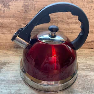 Puede incluir: Una tetera de acero inoxidable roja con un asa negra. La tetera tiene un acabado brillante y un borde plateado alrededor de la base.