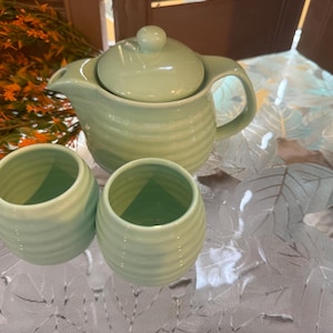 Könnte beinhalten: Eine hellgrüne Teekanne aus Keramik mit passendem Deckel und zwei Tassen. Die Teekanne und die Tassen haben ein geripptes Design.