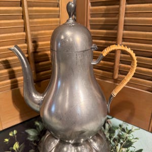 Może przedstawiać: Zabytkowy srebrny metalowy czajnik z zakrzywionym dziobkiem i plecionym uchwytem. Czajnik ma pękaty korpus i pokrywkę z małym pokrętłem. Stoi na dekoracyjnej podstawie. Tło przedstawia drewnianą okiennicę.