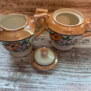 Puede incluir: Un juego de té vintage con base de color crema y melocotón y diseños florales. El juego incluye una tetera, una jarra de crema y un azucarero.