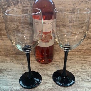 May include: Two black stemmed wine glasses with a bottle of pink wine in the background. The wine bottle label says "Winking Owl White Zinfandel".