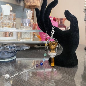 May include: A silver chain with a tiger's eye pendulum featuring an angel design. The pendulum is surrounded by seven chakra colored beads.