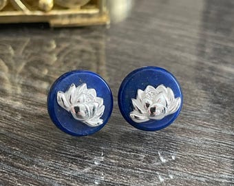 May include: A pair of round, dark blue stud earrings. Each earring features a silver lotus flower design. The earrings are displayed on a dark wooden surface, with a gold-colored decorative box in the background.