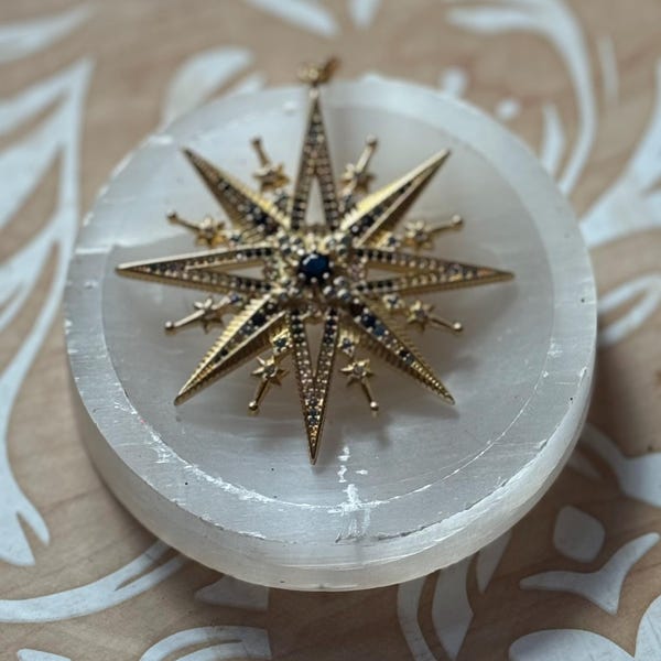 Puede incluir: Un colgante dorado en forma de estrella con detalles en negro y azul, colocado sobre una piedra blanca y circular. El colgante tiene una piedra central azul oscuro y está rodeado de pequeños detalles en forma de estrella. Probablemente sea una joya.