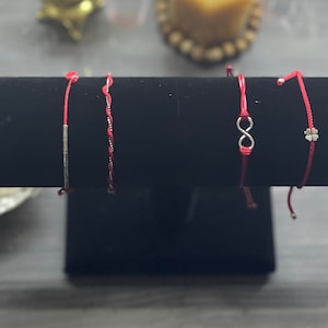 May include: Four red string bracelets with silver charms. The charms include a straight line, a braided design, an infinity symbol, and a four-leaf clover.