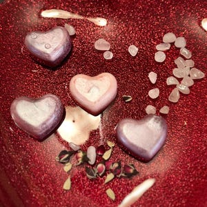 May include: A close-up of a red, glittery surface with four heart-shaped objects. Three hearts are a metallic purple color, and one is pink. Small, clear stones and dried rose petals are scattered around the hearts.