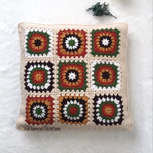 Puede incluir: Una almohada cuadrada de crochet con un patr&oacute;n geom&eacute;trico marr&oacute;n, blanco, verde, naranja y rojo. La almohada est&aacute; sobre una superficie blanca.
