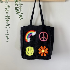 Op de afbeelding: Zwarte gehaakte tote bag met kleurrijke patches. De patches omvatten een regenboog, een vredesteken, een smiley en een bloem.