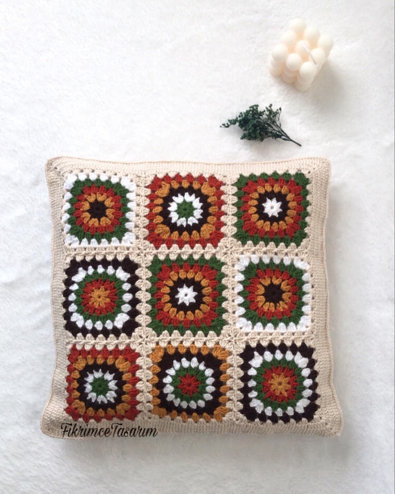 Puede incluir: Una almohada cuadrada de crochet con un patr&oacute;n geom&eacute;trico marr&oacute;n, blanco, verde, naranja y rojo. La almohada est&aacute; sobre una superficie blanca.