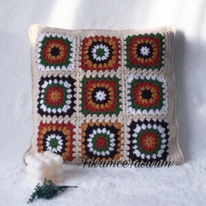 Puede incluir: Una almohada cuadrada de ganchillo con un patr&oacute;n geom&eacute;trico en tonos marrones, naranjas, verdes y blancos. La almohada est&aacute; hecha de cuadrados de la abuela y tiene un borde beige.