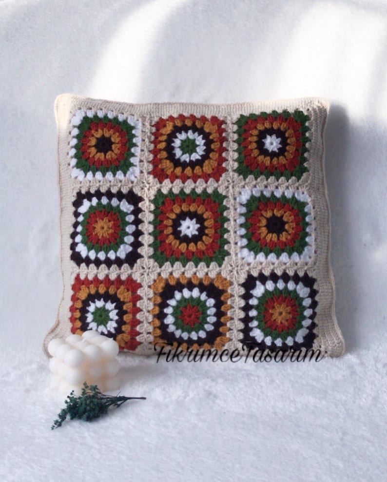 Puede incluir: Una almohada cuadrada de ganchillo con un patr&oacute;n geom&eacute;trico en tonos marrones, naranjas, verdes y blancos. La almohada est&aacute; hecha de cuadrados de la abuela y tiene un borde beige.