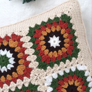 Puede incluir: Una almohada tejida a crochet con un patr&oacute;n cuadrado en tonos de marr&oacute;n, naranja, rojo, verde y blanco. La almohada est&aacute; hecha de hilo beige y tiene un borde simple.