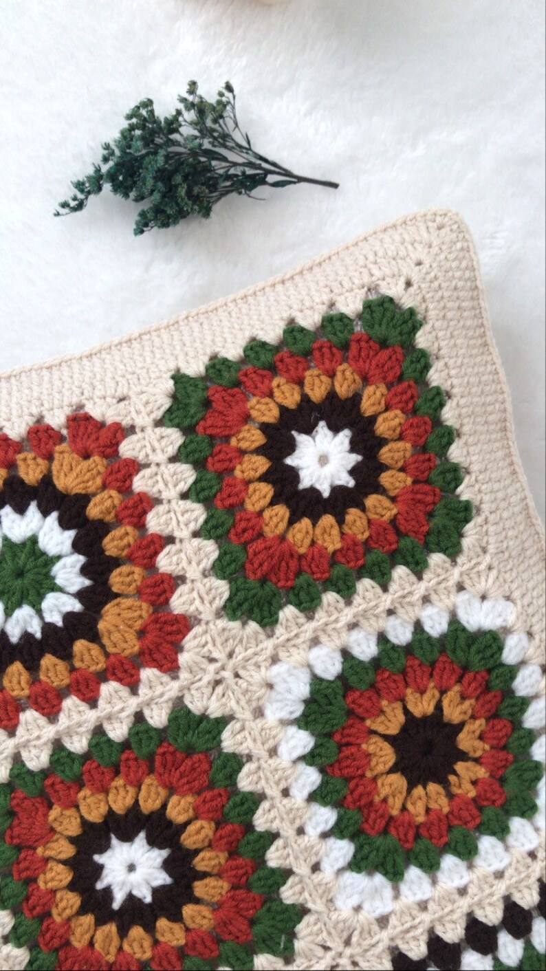Puede incluir: Una almohada tejida a crochet con un patr&oacute;n cuadrado en tonos de marr&oacute;n, naranja, rojo, verde y blanco. La almohada est&aacute; hecha de hilo beige y tiene un borde simple.