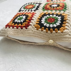 Puede incluir: Una almohada de crochet con fondo beige y patrones geom&eacute;tricos coloridos en tonos marrones, naranjas, verdes y blancos.