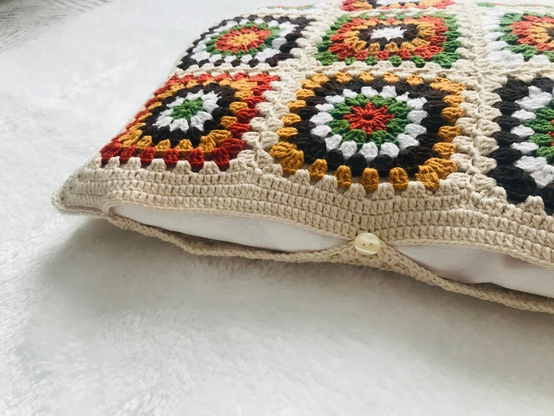 Puede incluir: Una almohada de crochet con fondo beige y patrones geom&eacute;tricos coloridos en tonos marrones, naranjas, verdes y blancos.